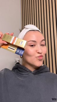 Huidverzorgingsproducten in de hand met goudkleurige en blauwe verpakkingen. De achtergrond bestaat uit verticale houten latten. Model draagt een grijze hoodie en een hoofdband.