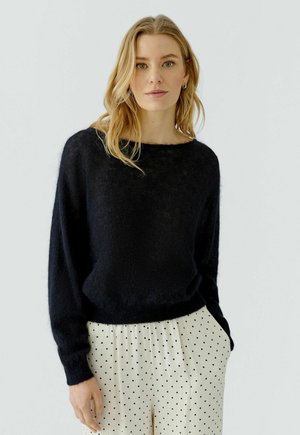 Vrouw met blond haar die een zwarte pluizige trui en witte broek met stippen draagt, staat tegen een effen achtergrond.