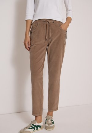 Femme portant un pantalon marron clair à cordon avec poches zippées, un haut blanc à manches longues, et des baskets décontractées beige et vertes, debout contre un mur uni.