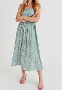 Femme aux longs cheveux blonds portant une robe midi verte à fleurs sans manches et des sandales à talons roses à brides, bras croisés.