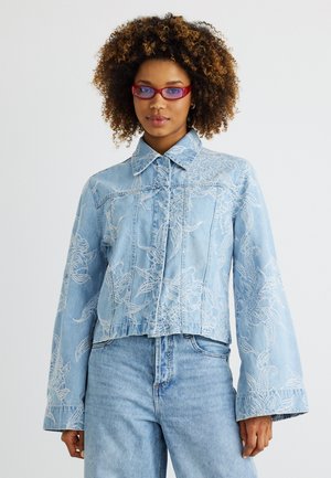 Mladá žena s kučeravými vlasmi nosí červené slnečné okuliare a svetlomodrú rifľovú bundu s bielou kvetinovou výšivkou, ktorú dopĺňajú modré džínsy.