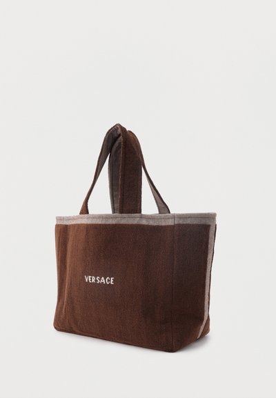 Καφέ ορθογώνια τσάντα ώμου με κοντά χερούλια και ανοιχτόχρωμη επένδυση, με λευκό κείμενο «VERSACE» στο κέντρο της μπροστινής πλευράς.