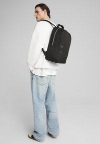 Zaino nero con un design strutturato, maniglia superiore e spallacci regolabili. Presenta una tasca frontale e una cerniera argentata per un tocco di stile.