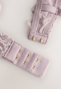 Nėrinių ir audinio liemenėlės dirželis švelnios alyvinės spalvos su auksinėmis detalėmis ir kabliukų užsegimu, pasižymintis tekstūruotais siūtais ir banguotais kraštais.