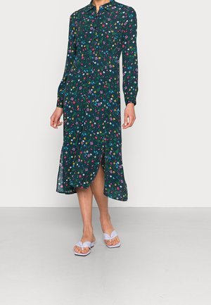 Vrouw draagt een donkergroene midi-jurk met bloemenprint, knoopsluiting, lange mouwen en een voorzijde met een split, gecombineerd met witte sandalen met hak.