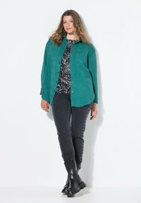 Turkusowa zamszowa koszula, czarna wzorzysta bluzka, ciemne jeansy ze srebrnymi ćwiekami oraz czarne masywne buty. Model stoi na tle białym.