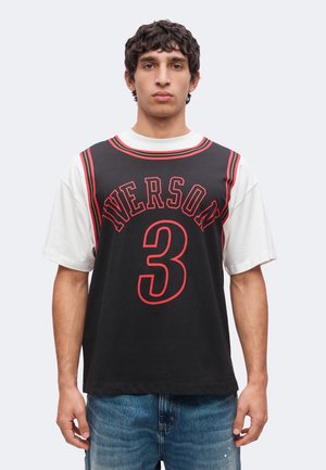 Ung mand med krøllet mørkt hår iført en sort og rød basketballtrøje over en hvid t-shirt og blå jeans, stående og vendt fremad.