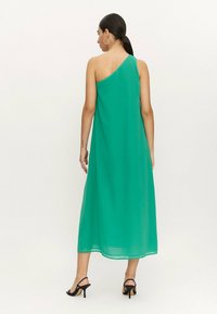 Vestido maxi verde de un solo hombro hecho de tejido ligero. Presenta un diseño fluido y un dobladillo que llega a la mitad de la pantorrilla, combinado con tacones negros.