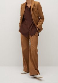 Blazer marron sur une chemise marron à boutons, associé à un pantalon large assorti et des chaussures claires. Texture de tissu lisse.