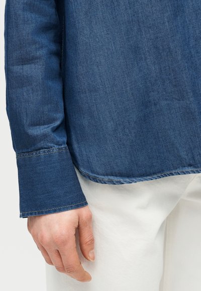 Ανδρικό χέρι που ξεκουράζεται στο πλάι, φοράει μακρυμάνικο μπλε τζιν πουκάμισο και λευκό παντελόνι, δείχνοντας την μανσέτα του πουκαμίσου και την υφή του υφάσματος.