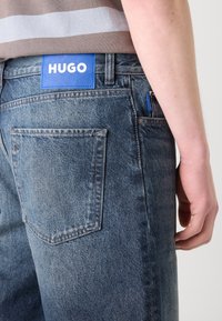 Blå denimjeans med en falmet finish, med et blåt mærke med "HUGO" på taljebåndet og to baglommer.