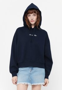 Marineblå cropped hettegenser med snorhette og brodert logo. Mykt bomullsmateriale og ribbede mansjetter. Kombinert med et slitt denimskjørt.