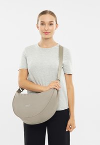 Šedá tkaninová crossbody taška so zaobleným tvarom, nastaviteľným popruhom a jemným brandingom. Mäkká textura, minimalistický dizajn.