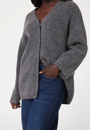 Femme portant un cardigan gris ample et doux à manches longues sur un jean bleu foncé, une main dans la poche, devant un fond uni.