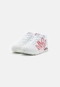 Vita Skechers sneakers med ljusblå snören och röd graffiti-inspirerad design på sidorna, visade från en lätt vinkel på en vit bakgrund.