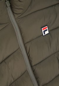 Vadderad väst i mörk olivgrön med en central dragkedja och diagonalt kviltad textur. Har en liten broderad logotyp i röd, vit och blå.