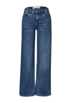 Personne portant un jean denim bleu foncé et un haut blanc, tenant un sac à main en daim marron avec des bagues dorées aux doigts.