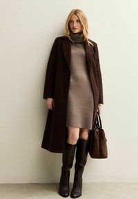 Een bruine lange jas over een beige gebreide jurk, kniehoge bruine laarzen en een donkerbruine handtas. Gladde texturen met minimale patronen.