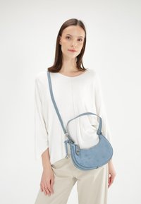 Blauwe suède schoudertas met een gebogen vorm, verstelbare band en zilverkleurige hardware accenten. Model draagt een witte top en beige broek.