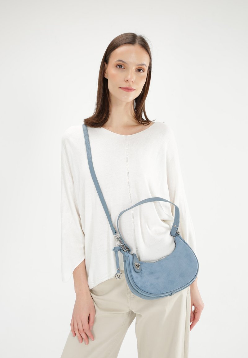 Blauwe suède schoudertas met een gebogen vorm, verstelbare band en zilverkleurige hardware accenten. Model draagt een witte top en beige broek.