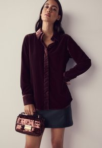 Bordeauxrotes Samthemd mit Kragen und Knopfleiste, kombiniert mit einem schwarzen Kunstlederrock. Hält eine strukturierte schwarz-pinke Handtasche.