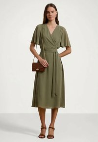 Mulher vestindo um vestido envolvente verde oliva com mangas flutuantes, bolsa de ombro castanha e sandálias de tiras no tornozelo castanhas combinando, posando contra um fundo liso.