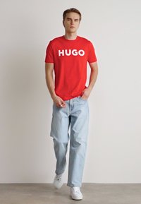 Camiseta de algodón roja con "HUGO" impreso en letras grandes blancas, combinada con jeans de mezclilla azul claro y zapatillas blancas. Ajuste casual.