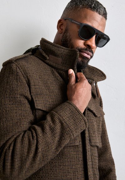 Veste à carreaux marron avec un col haut et des poches à boutons, associée à des lunettes de soleil noires avec des accents texturés.