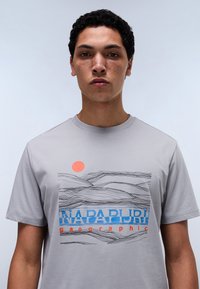 T-shirt en coton gris à manches courtes, présentant un design graphique de paysage, un soleil orange et le texte "NAPAPIJRI geographic" en bleu et rouge.