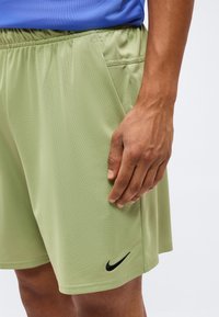 Άτομο που φοράει ανοιχτό πράσινο σορτς Nike με μαύρο λογότυπο στο αριστερό πόδι και μπλε μπλούζα, χέρι ακουμπισμένο δίπλα στην τσέπη του σορτς.