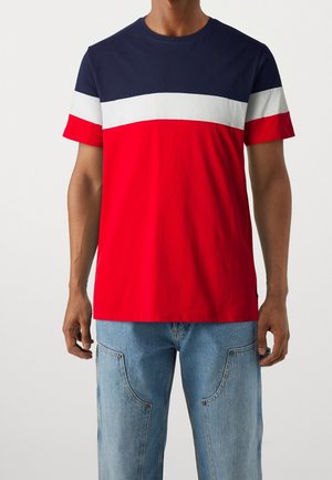 Mand iført en kortærmet t-shirt med horisontale farveblokke i marineblå, hvid og rød, kombineret med lyseblå jeans.