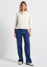 Camicia beige chiaro con chiusura a bottoni e ricami ad occhio sulla parte inferiore, abbinata a jeans a gamba larga blu e scarpe di colore chiaro.