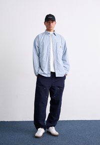 Lys blå stribet skjorte over en hvid t-shirt, parret med mørk marineblå vide bukser og hvide sneakers. Sort baseballkasket inkluderet.