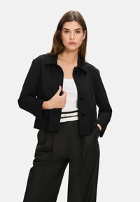 Black cropped jacket with a collar, front buttons, and flap pockets, paired with a white top and black high-waisted trousers.