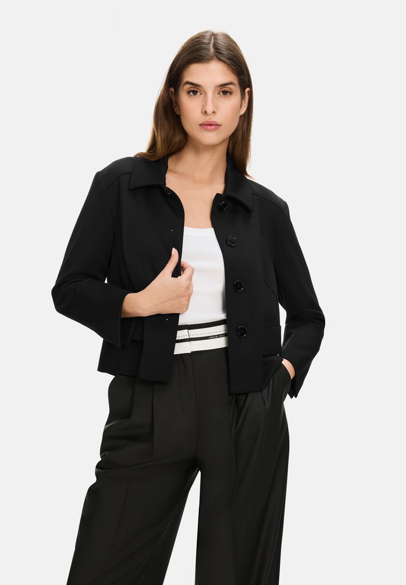 Black cropped jacket with a collar, front buttons, and flap pockets, paired with a white top and black high-waisted trousers.