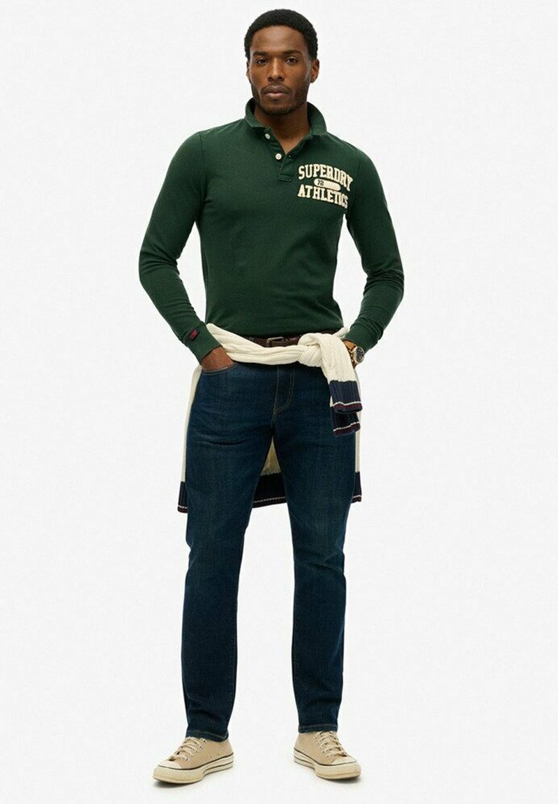Groene langemouwen polo met de tekst "Superdry Athletics", gecombineerd met donkerblauwe jeans en beige sneakers, een witte trui om de taille geknoopt.