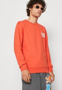 Felpa arancione con collo rotondo e polsini a costine, con un logo bianco "Tommy Jeans" sul lato sinistro del petto. Indossata con pantaloni corti grigi.