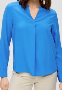 Blouse bleu à manches longues avec un col en V, présentant un pli discret à l'avant. Fabriquée en tissu lisse, avec une coupe décontractée et un ourlet droit.