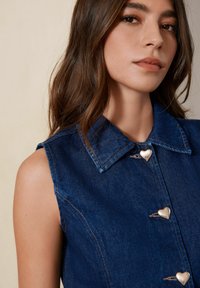 Denim bezrukavni top s ovratnikom i dugmadi u obliku srca. Tkanina je tamnoplava s kontrastnim šavovima.