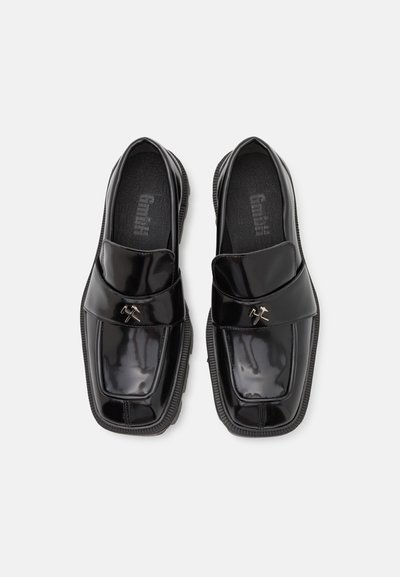 Black patent leather loafers with a square toe, featuring a wide strap and decorative metallic hardware. Textured black inner lining.
