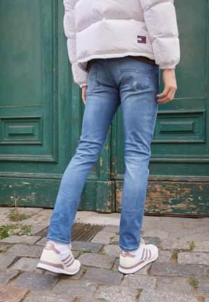 Personne portant un jean bleu clair, des baskets Adidas blanches et une doudoune blanche, debout sur des pavés devant une porte verte usée.