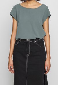 Donna che indossa un top verde chiaro a maniche corte infilato in una gonna di jeans nera a vita alta con cuciture bianche e uno spacco anteriore.