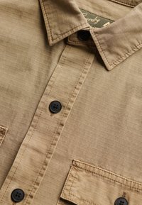 Beige overhemd met knopen, zwarte knopen, een puntige kraag en een rechthoekig zakje met stiksel op een gestructureerde stof.