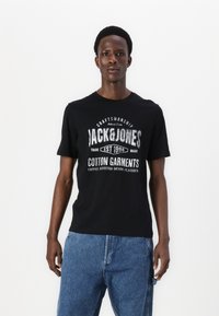 Black cotton t-shirt featuring a graphic print with "JACK & JONES" in white and silver accents, paired with light blue denim shorts.
