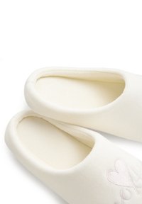 Witte stoffen slippers met een zachte binnenvoering, open achterkant ontwerp, en subtiele roze geborduurde hartdetail aan de zijkant.
