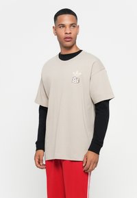Beige bomulds T-shirt med korte ærmer og rund hals, med et lille sort logo og lagdelt over en sort langærmet skjorte.