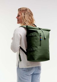Zelený batoh s rolovacím víkem vyrobený z voděodolného materiálu, s nastavitelnými popruhy, zipovou kapsou a kontrastními kovovými detaily.