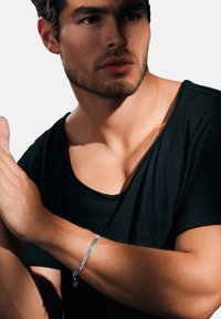 Silvrigt metallarmband med en texturerad yta, prytt med en berlock, bärs på ett bar handle. Modell i en svart t-shirt med öppen halsringning.