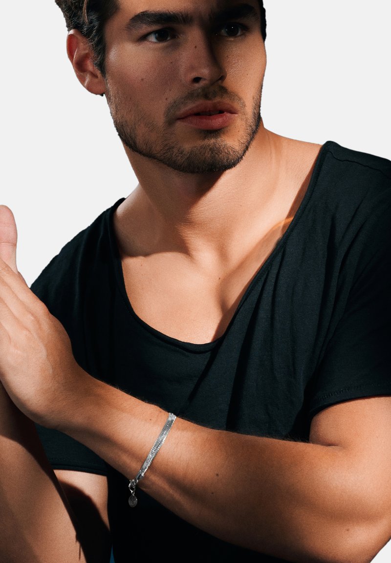 Silvrigt metallarmband med en texturerad yta, prytt med en berlock, bärs på ett bar handle. Modell i en svart t-shirt med öppen halsringning.