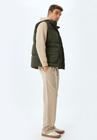 Grøn polstret vest, beige hættetrøje under, kremfarvede bukser, lysebrune sneakers. Funktioner inkluderer to frontlommer og en høj krave.